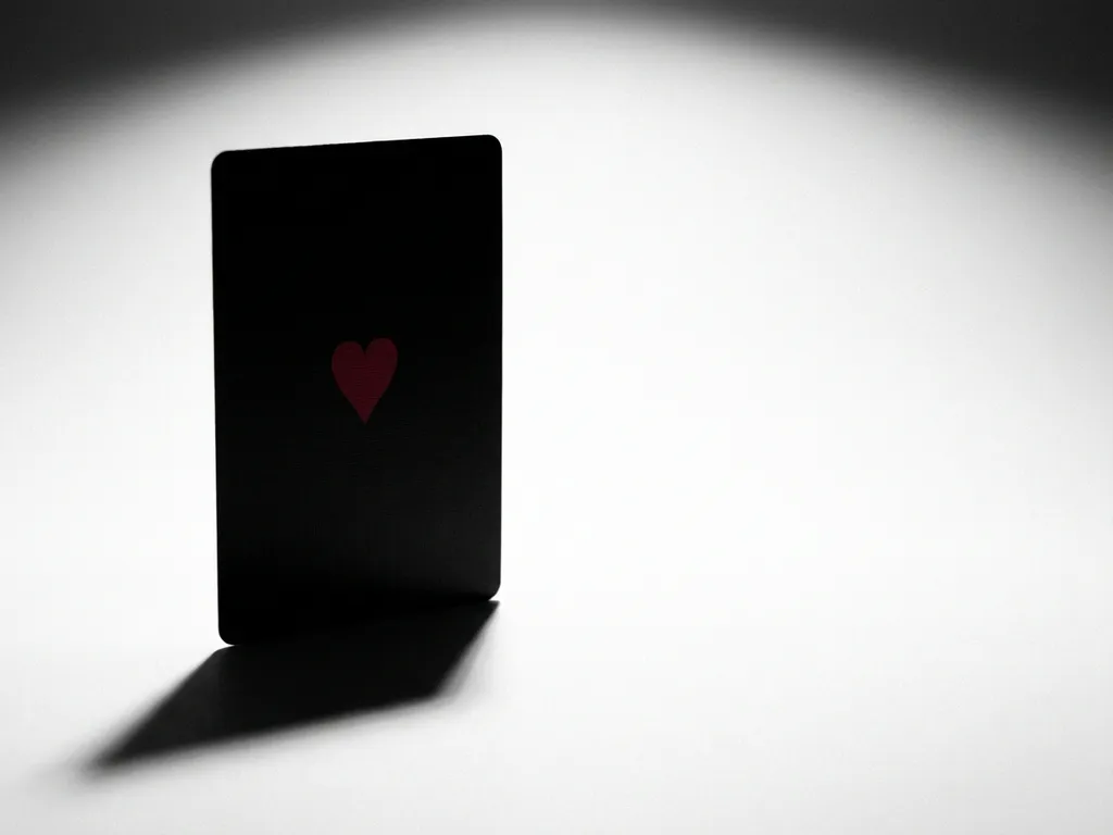 King of Hearts playing card standing upright, matte black back with crimson heart emblem, dramatic spotlight shadow on white surface.