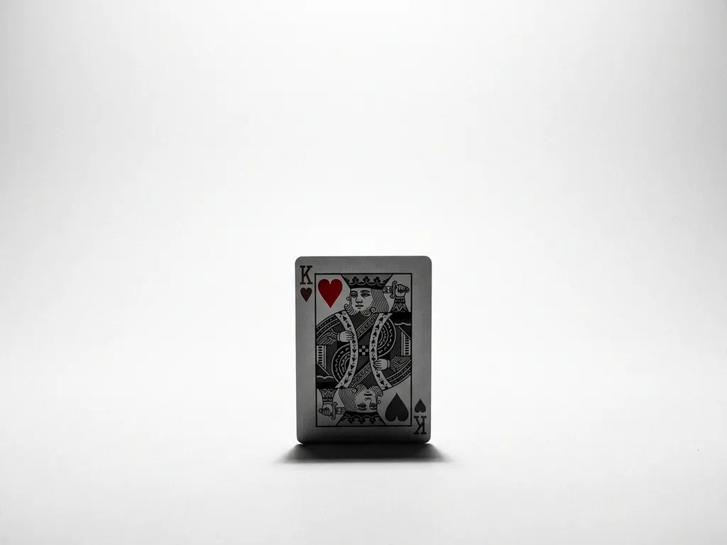 King of Hearts playing card standing upright on white surface, matte black tones with crimson heart suit accent, dramatic overhead studio lighting.
