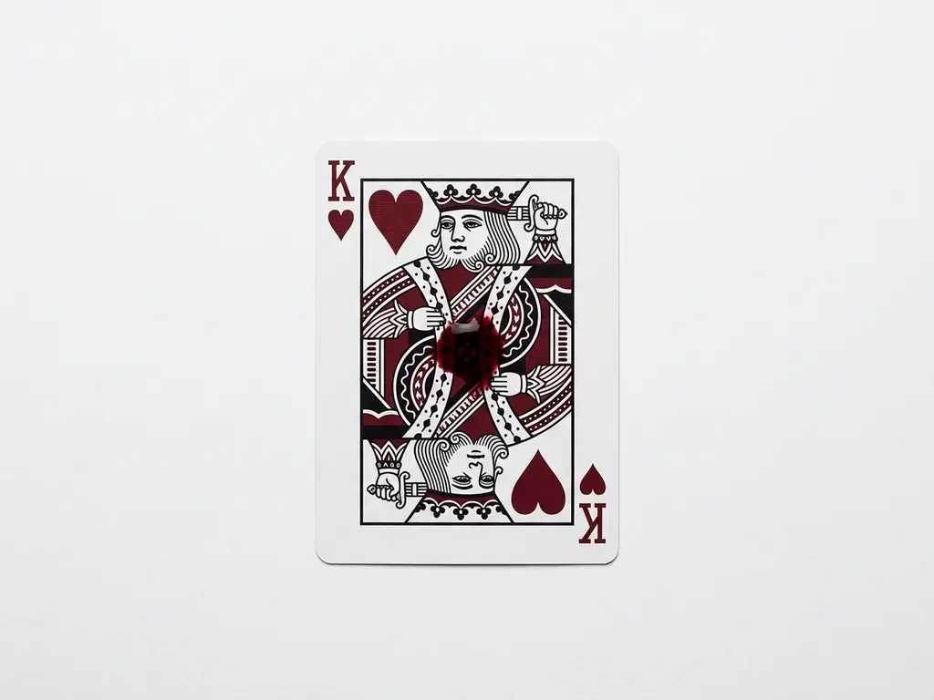 King of Hearts playing card lying flat on white surface with a crimson ink droplet bleeding from its center.
