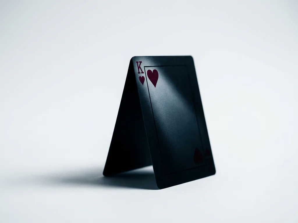 Matte black King of Hearts playing card standing upright, crimson heart symbol lit by directional studio light against a white background.