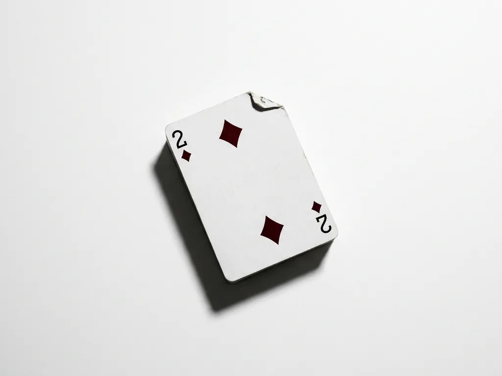 Two of diamonds playing card standing upright on white surface, slightly warped with dog-eared corner, studio lighting casting precise shadow.